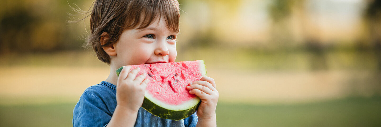 Kleines Mädchen im Jeanskleid isst ein Stück gesunde Wassermelone.