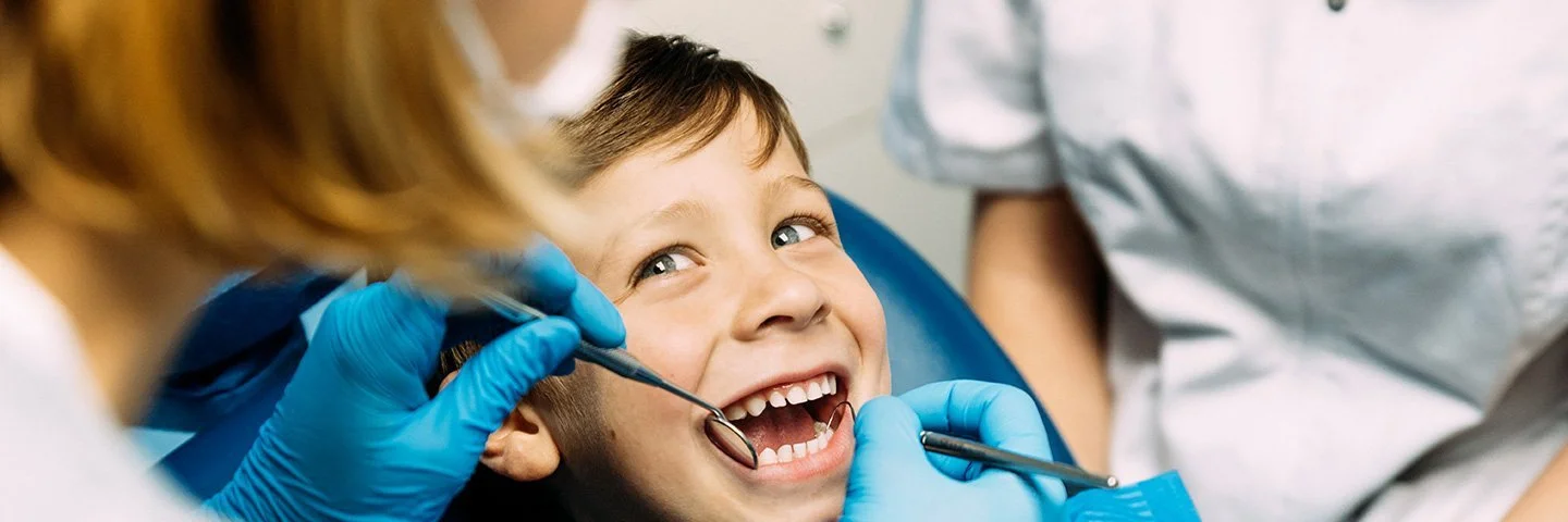 Ein kleiner Junge liegt lächelnd auf einem blauen Behandlungsstuhl, während eine Zahnärztin mit blauen Handschuhen seine Zähne untersucht.