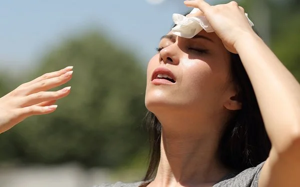 Eine Frau steht bei Sonnenschein im Freien. Sie kühlt ihre Stirn mit einem Tuch und fächelt sich Luft zu.