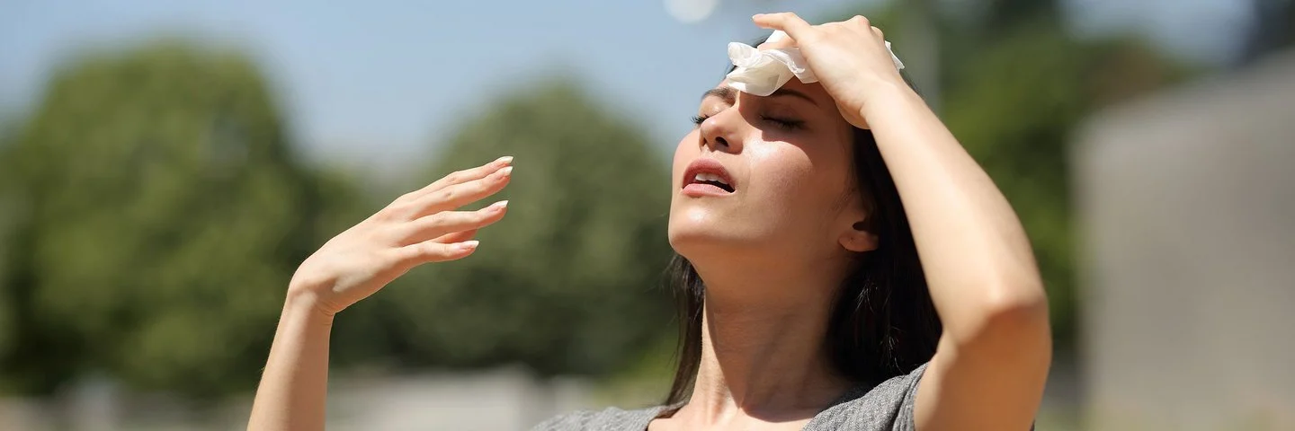 Eine Frau steht bei Sonnenschein im Freien. Sie kühlt ihre Stirn mit einem Tuch und fächelt sich Luft zu.