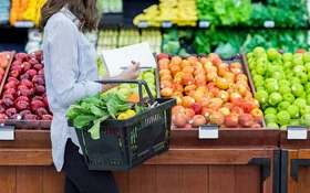 Eine Frau mit Einkaufskorb und Einkaufszettel kauft in einem Supermarkt Obst ein und sucht nach Obst mit wenig Zucker..