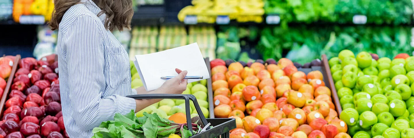 Eine Frau mit Einkaufskorb und Einkaufszettel kauft in einem Supermarkt Obst ein und sucht nach Obst mit wenig Zucker..