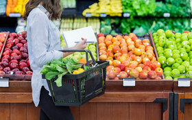 Eine Frau mit Einkaufskorb und Einkaufszettel kauft in einem Supermarkt Obst ein und sucht nach Obst mit wenig Zucker..