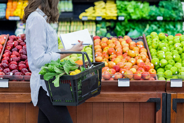 Eine Frau mit Einkaufskorb und Einkaufszettel kauft in einem Supermarkt Obst ein und sucht nach Obst mit wenig Zucker..
