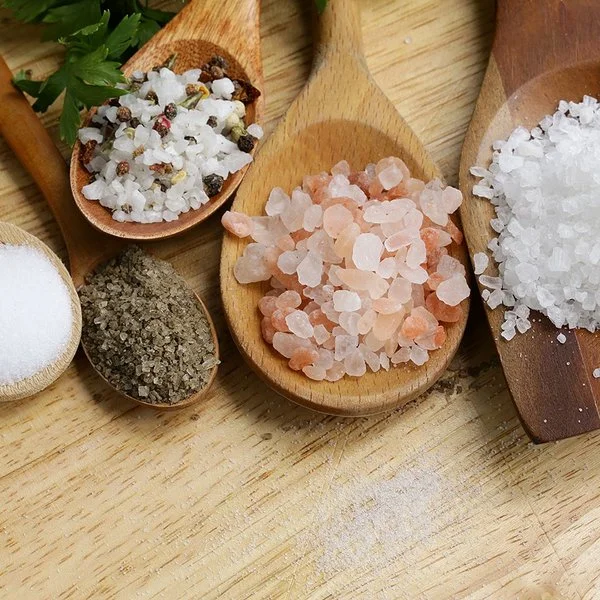Fünf Holzlöffel mit verschiedenen Salzarten liegen auf einem Tisch.