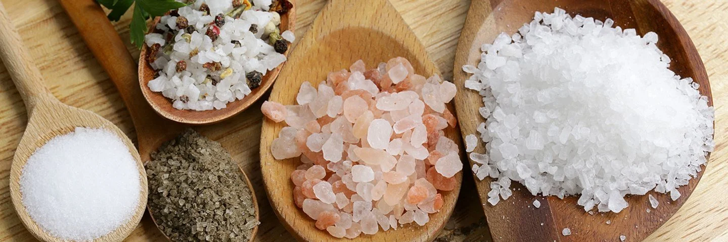 Fünf Holzlöffel mit verschiedenen Salzarten liegen auf einem Tisch.