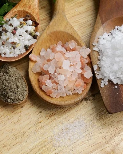 Fünf Holzlöffel mit verschiedenen Salzarten liegen auf einem Tisch.