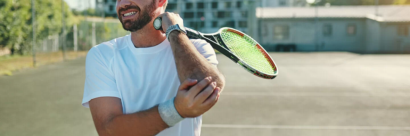 Ein Mann auf dem Tennisplatz fasst sich mit schmerzerfülltem Gesicht an seinen Tennisarm.