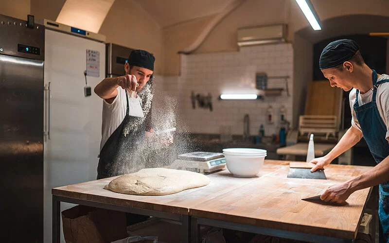 In einer Backstube arbeiten zwei Bäcker an einem Holztisch. Der eine bestäubt einen großen Teigfladen mit Mehl, der andere schiebt mit Spachteln Mehlreste zusammen.