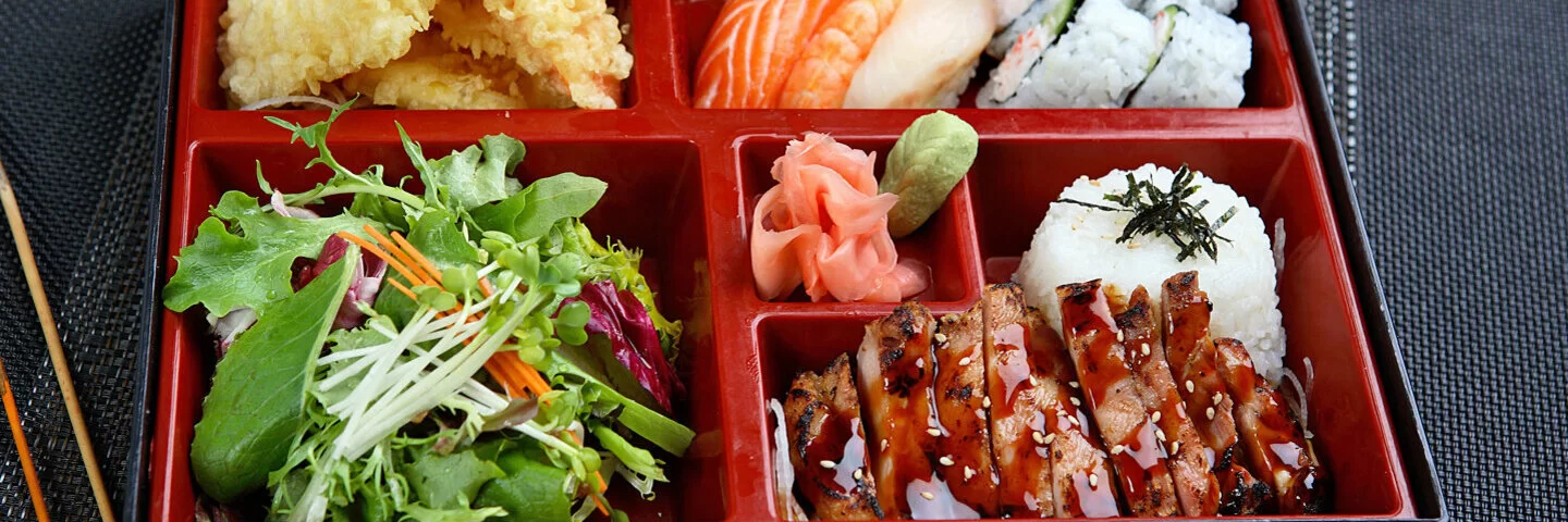 Klassische japanische Bento-Box mit 5 unterschiedlich großen Fächern mit Sushi, Teriyaki-Huhn, Tempura-Shrimps und Salat.