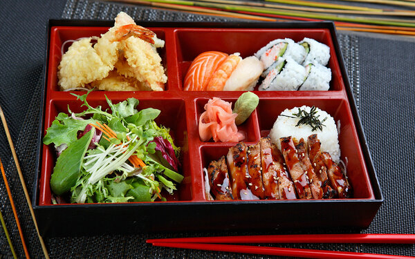 Klassische japanische Bento-Box mit 5 unterschiedlich großen Fächern mit Sushi, Teriyaki-Huhn, Tempura-Shrimps und Salat.
