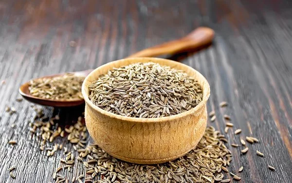 Auf einem Holztisch steht ein Holzschälchen mit Kreuzkümmelsamen. Hinter dem Schälchen liegt ein Holzlöffel, ebenfalls mit Kreuzkümmelsamen. Weitere Samen sind um das Schälchen herum auf dem Tisch verstreut.