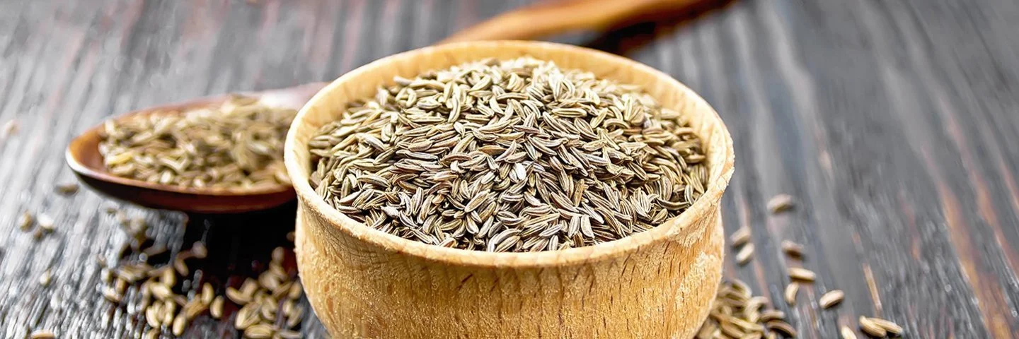 Auf einem Holztisch steht ein Holzschälchen mit Kreuzkümmelsamen. Hinter dem Schälchen liegt ein Holzlöffel, ebenfalls mit Kreuzkümmelsamen. Weitere Samen sind um das Schälchen herum auf dem Tisch verstreut.