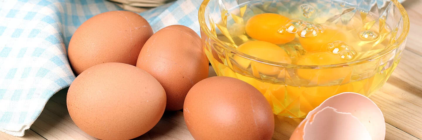 Braune Eier liegen auf einem hölzernen Untergrund. Daneben steht eine Schüssel mit aufgeschlagenen Eiern.