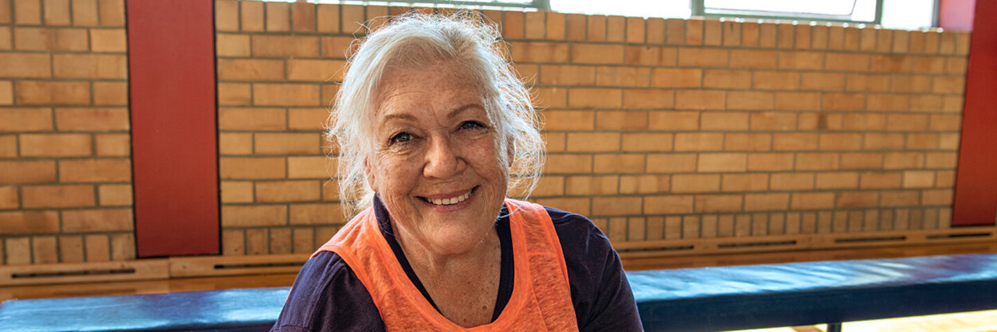Eine ältere Frau sitzt in einer Turnhalle und schaut lächelnd in die Kamera. Die Faktenboxen der AOK bieten Versicherten Informationen um viele Gesundheitsthemen.