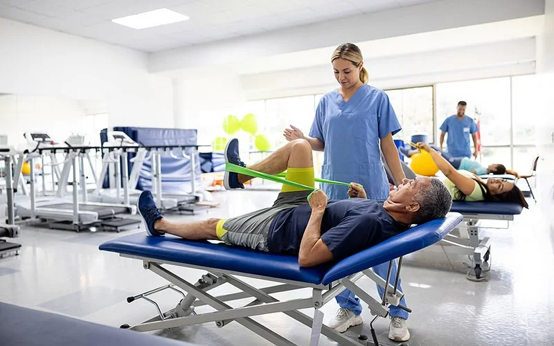 Ein Mann bei der Physiotherapie macht unter Anleitung der Therapeutin Übungen mit einem Stretchband.