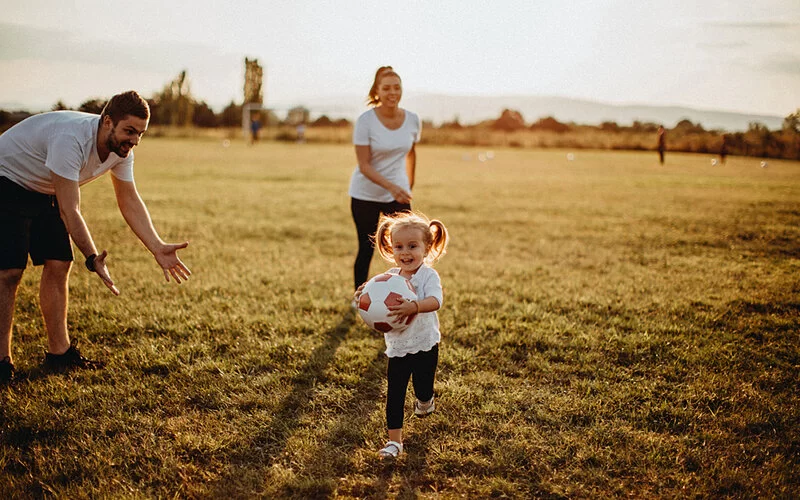 Mutter, Vater und ihre kleine Tochter spielen mit einem Fußball auf einer Wiese.