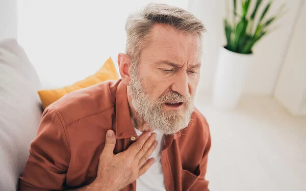 Ein älterer Mann mit angestrengtem Gesichtsausdruck sitzt auf einem Sofa und legt die rechte Hand an den oberen Teil seiner Brust.