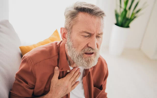 Ein älterer Mann mit angestrengtem Gesichtsausdruck sitzt auf einem Sofa und legt die rechte Hand an den oberen Teil seiner Brust.