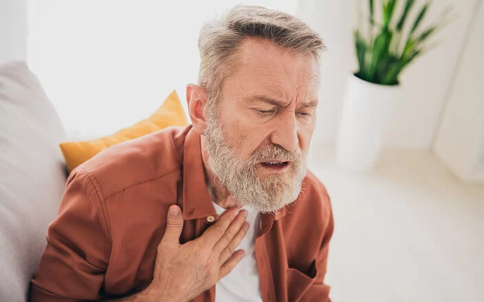 Ein älterer Mann mit angestrengtem Gesichtsausdruck sitzt auf einem Sofa und legt die rechte Hand an den oberen Teil seiner Brust.