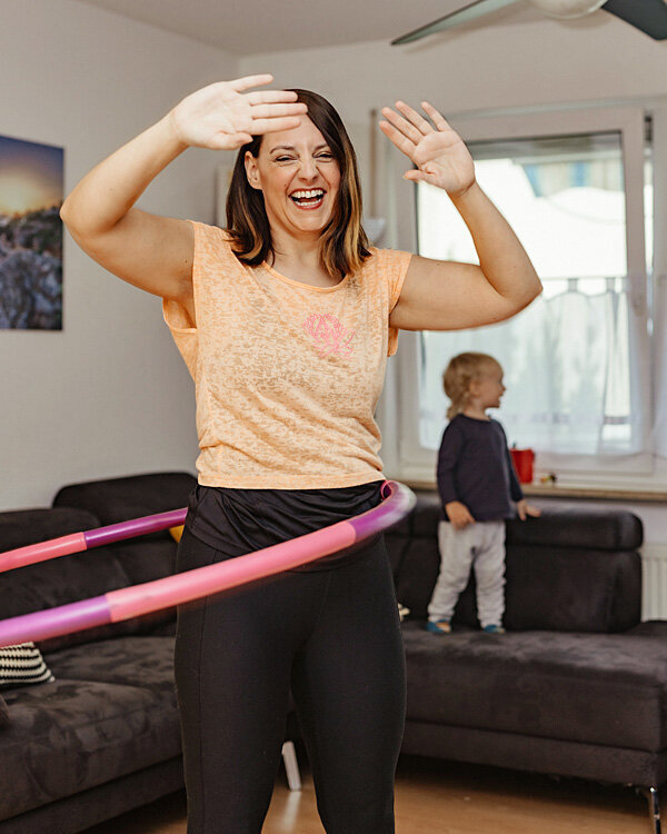 Alexandra trainiert im Wohnzimmer mit einem Hula-Hoop, ihr Kind steht im Hintergrund auf der Couch.