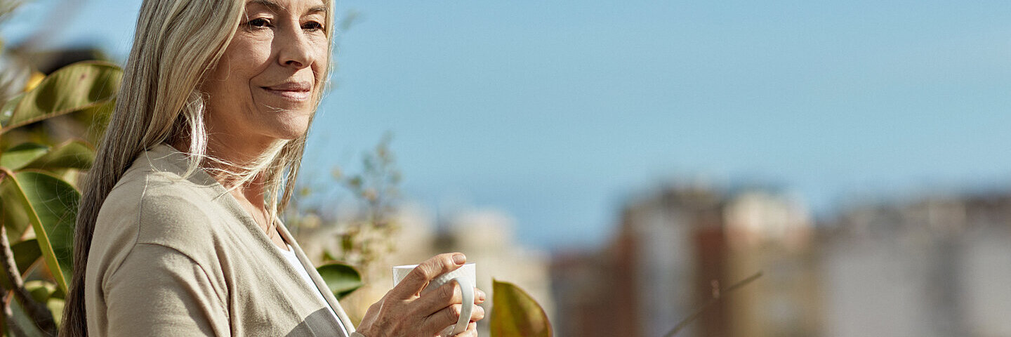 Eine ältere Frau nimmt sich zur Stressbewältigung eine Auszeit, mit einer Tasse Kaffee in der Natur.