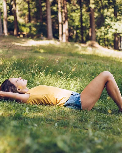 Junge Frau in gelbem T-Shirt und Jeans-Shorts, die in einer schattigen Waldlichtung im Gras liegt.