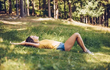 Junge Frau in gelbem T-Shirt und Jeans-Shorts, die in einer schattigen Waldlichtung im Gras liegt.
