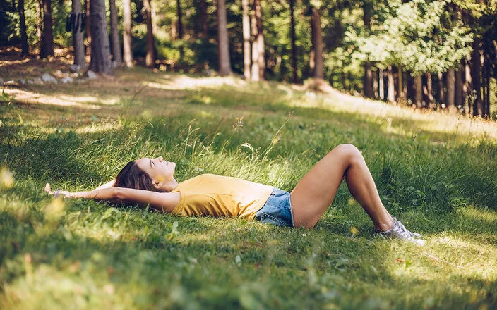 Junge Frau in gelbem T-Shirt und Jeans-Shorts, die in einer schattigen Waldlichtung im Gras liegt.