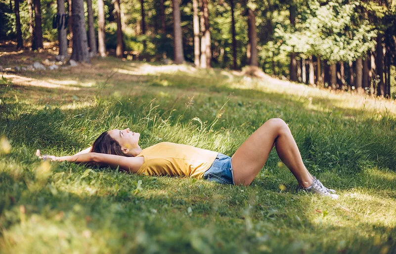 Junge Frau in gelbem T-Shirt und Jeans-Shorts, die in einer schattigen Waldlichtung im Gras liegt.