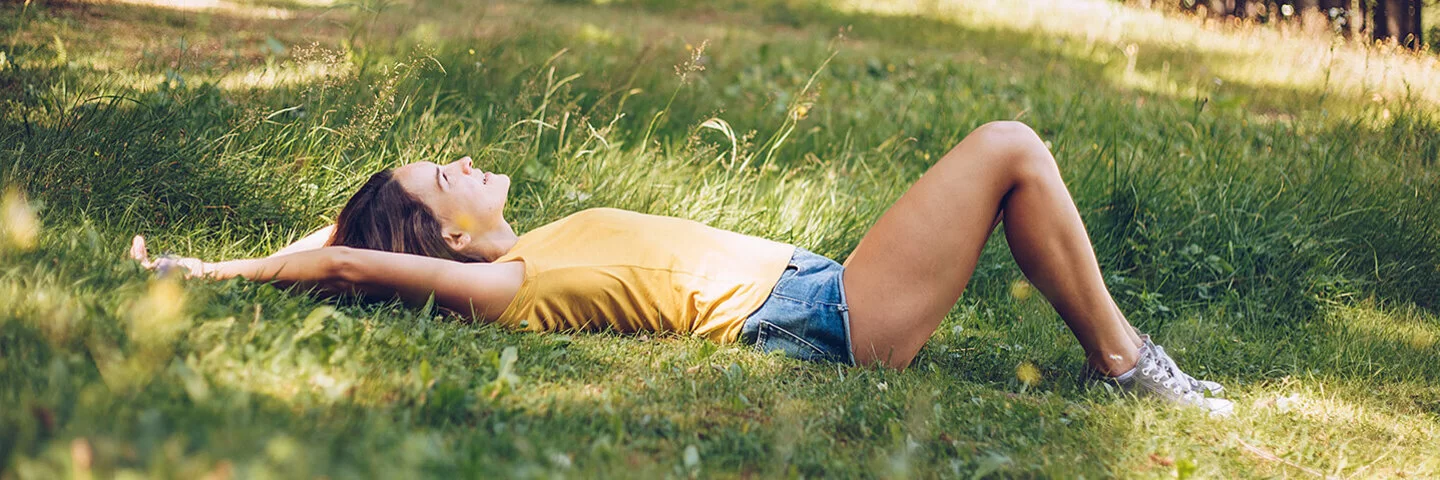 Junge Frau in gelbem T-Shirt und Jeans-Shorts, die in einer schattigen Waldlichtung im Gras liegt.