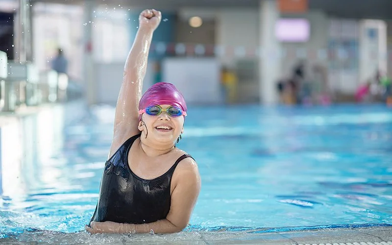 Ein Mädchen mit Übergewicht streckt sich mit dem Arm nach oben aus dem Schwimmbecken.