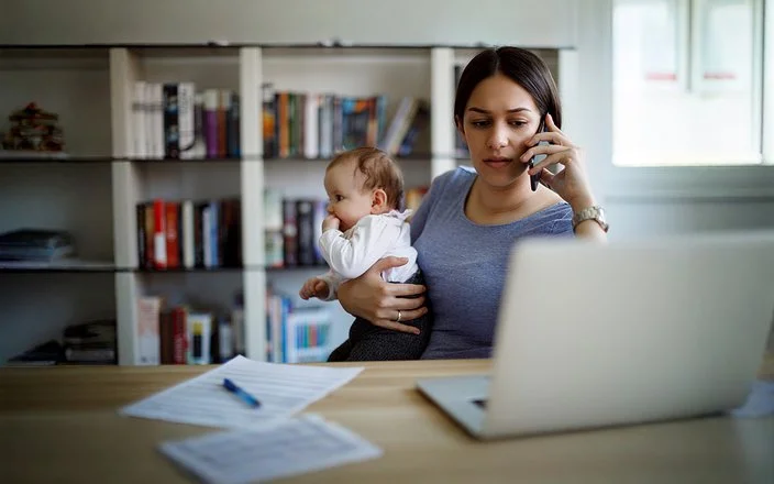 Eine junge Mutter hält ein Baby auf dem Arm, telefoniert gleichzeitig und sitzt vor einem Laptop mit Unterlage – Symbolbild für Mental Load im Familienalltag.