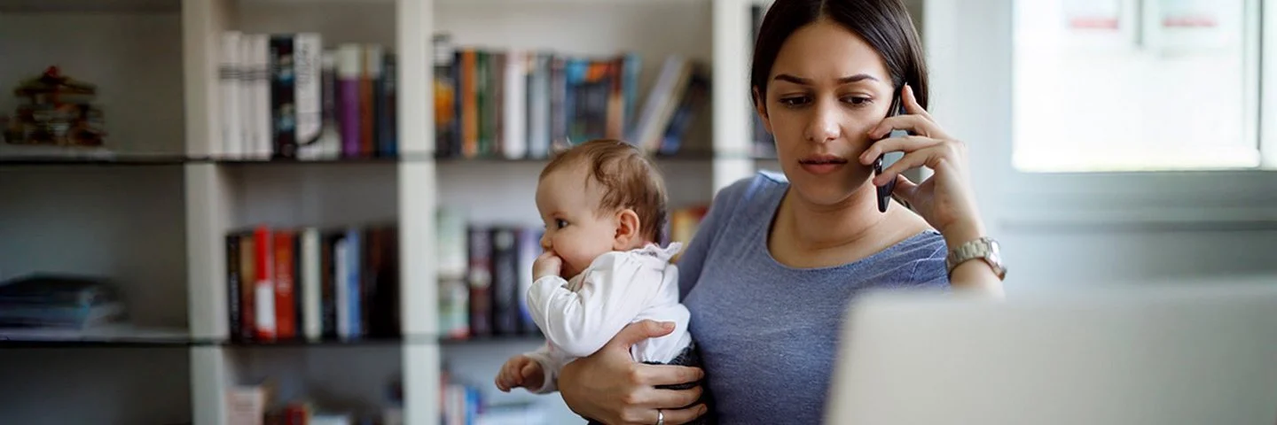 Eine junge Mutter hält ein Baby auf dem Arm, telefoniert gleichzeitig und sitzt vor einem Laptop mit Unterlage – Symbolbild für Mental Load im Familienalltag.
