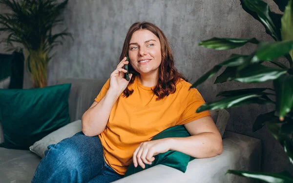 Eine leicht mehrgewichtige Frau Mitte 30 in legerer Kleidung sitzt entspannt auf einem von Grünpflanzen umrahmten Sofa und telefoniert.