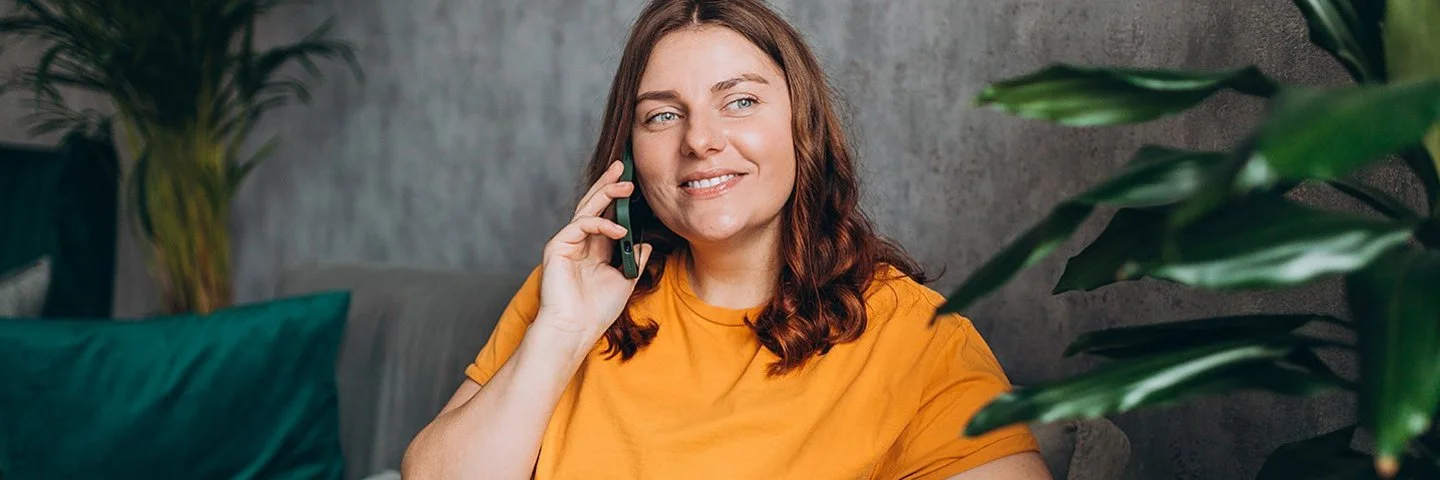 Eine leicht mehrgewichtige Frau Mitte 30 in legerer Kleidung sitzt entspannt auf einem von Grünpflanzen umrahmten Sofa und telefoniert.