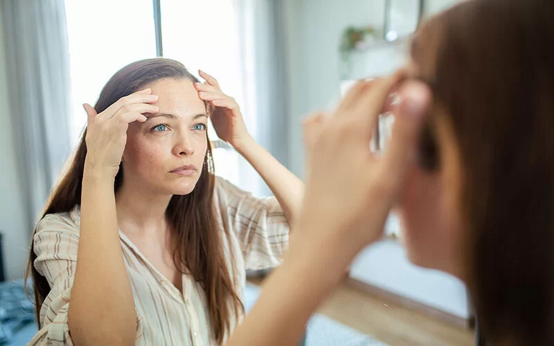 Eine Frau um die Dreißig, mit langen braunen Haaren, steht vor einem Spiegel und betrachtet kritisch ihre Stirn.