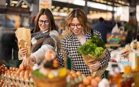 Zwei Frauen stehen vor einem Stand auf einem Lebensmittelmarkt und haben Papiertüten mit Gemüse in den Armen.