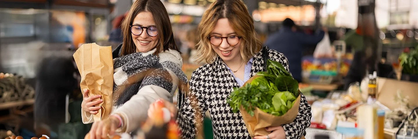 Zwei Frauen stehen vor einem Stand auf einem Lebensmittelmarkt und haben Papiertüten mit Gemüse in den Armen.