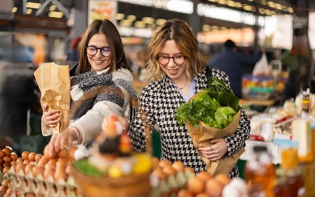 Zwei Frauen stehen vor einem Stand auf einem Lebensmittelmarkt und haben Papiertüten mit Gemüse in den Armen.
