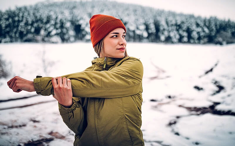 Eine junge Frau in winterlicher Kleidung dehnt sich in einer verschneiten Landschaft bei kalten Temperaturen, um nicht zu frieren.