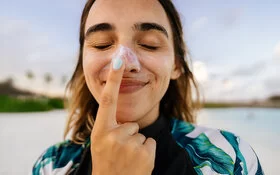 Eine junge Frau cremt sich die Nase mit Sonnencreme ein, um sich vor der Entstehung von einem Basaliom zu schützen.