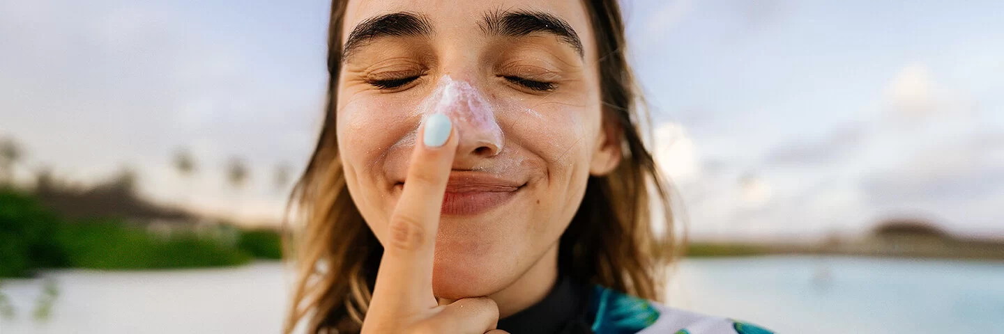 Eine junge Frau cremt sich die Nase mit Sonnencreme ein, um sich vor der Entstehung von einem Basaliom zu schützen.