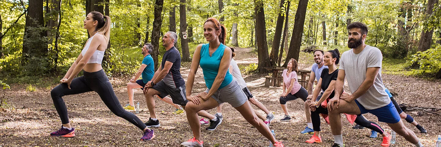 Eine Gruppe von Frauen und Männern in Sportkleidung dehnt ihren rechten Oberschenkel im Wald.