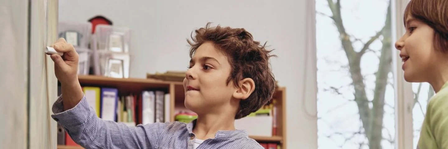 Zwei Kinder stehen vor einer Tafel und zeichnen mit Kreide; eines schreibt, das andere hält ein Kreidestück bereit. Im Hintergrund sitzt eine Lehrkraft an einem Tisch.