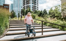 Eine ältere Frau mit einer Gehhilfe geht im urbanen Raum Treppen hinunter. Im Hintergrund sind Hochhäuser zu sehen. 