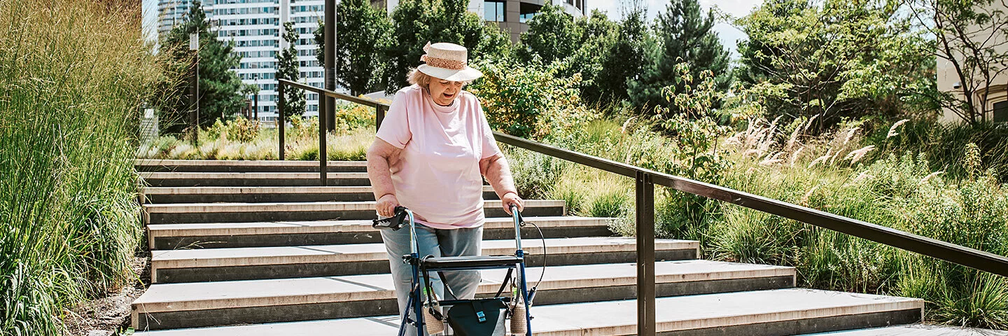 Eine ältere Frau mit einer Gehhilfe geht im urbanen Raum Treppen hinunter. Im Hintergrund sind Hochhäuser zu sehen.