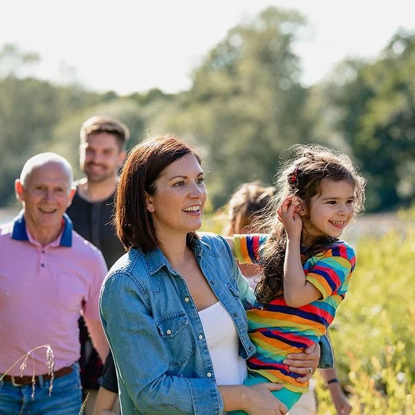 Eine Familie, der drei Generationen angehören, macht einen gemeinsamen Spaziergang. Sie stehen auf einem Feld, im Hintergrund schimmert Wassser, die Sonne scheint. Die Mutter hält ihre Tochter fest dem Arm. Beide lachen.