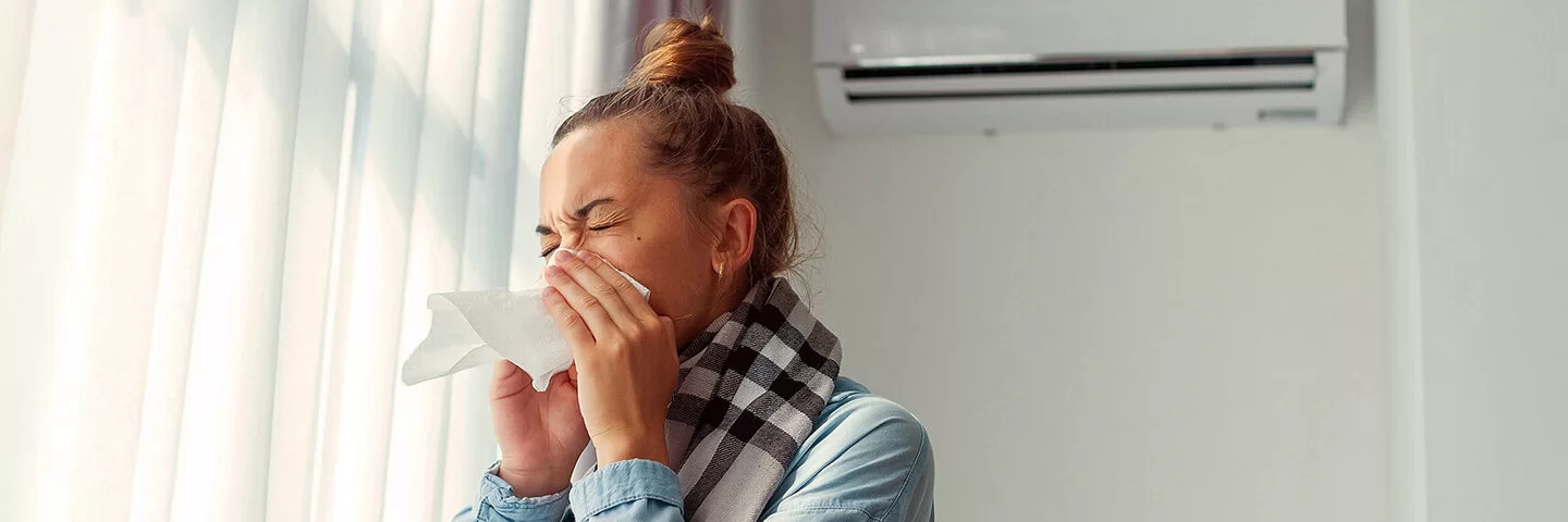 Niesende Frau, die ein Taschentuch vor ihr Gesicht hält, in einem hellen Raum mit zugezogenen Vorhängen und Klimaanlage an der Wand.