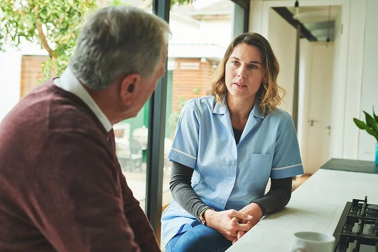 Eine Pflegeberaterin sitzt in einer Küche einem älteren Mann gegenüber. Sie unterhalten sich.